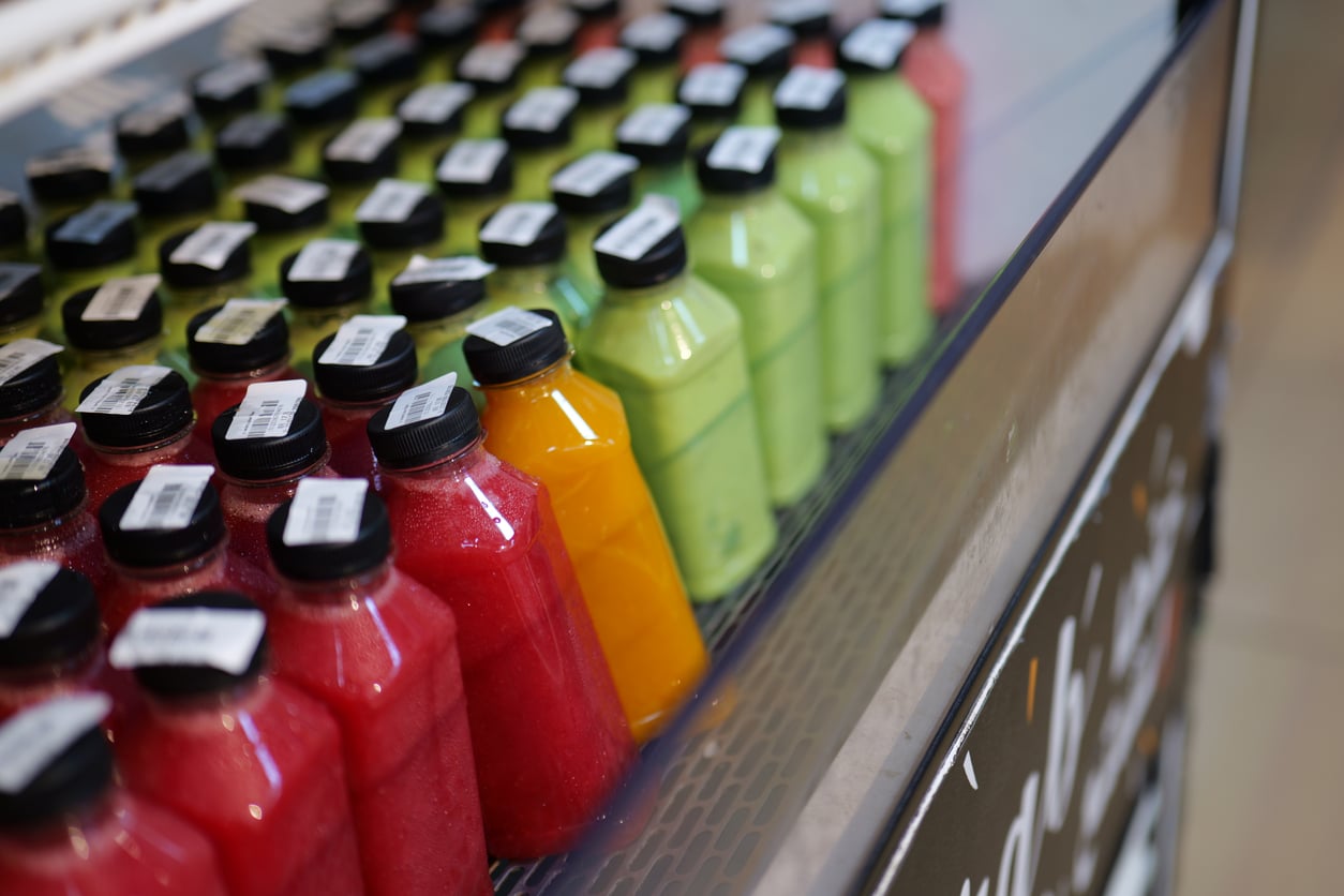Rows of plastic bottles filled with red, orange, and green juices are neatly arranged on a refrigerated display shelf, each bottle sealed with a black cap and a barcode sticker.