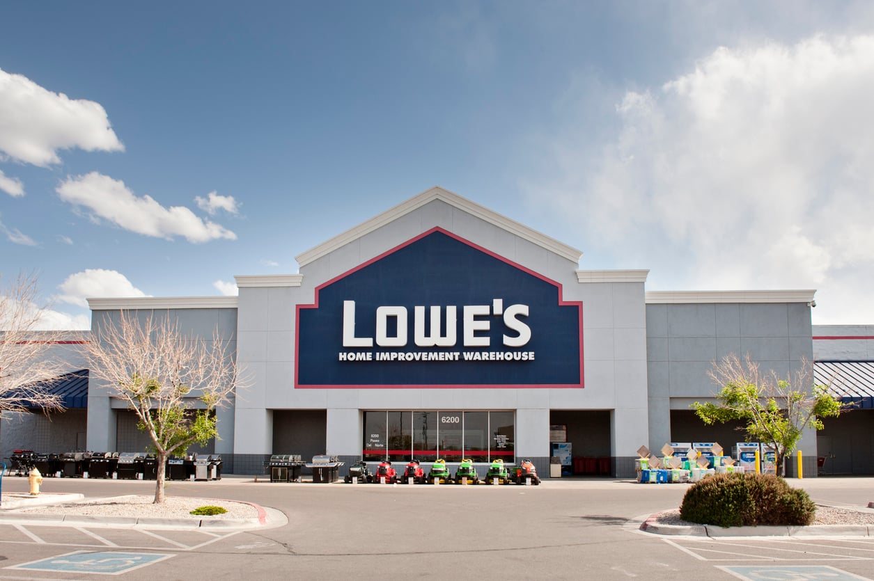 Front view of a Lowe’s Home Improvement Warehouse store with a large sign, blue and white exterior, clear sky, and various lawn equipment displayed outside the entrance amid news of 2026 layoffs.
