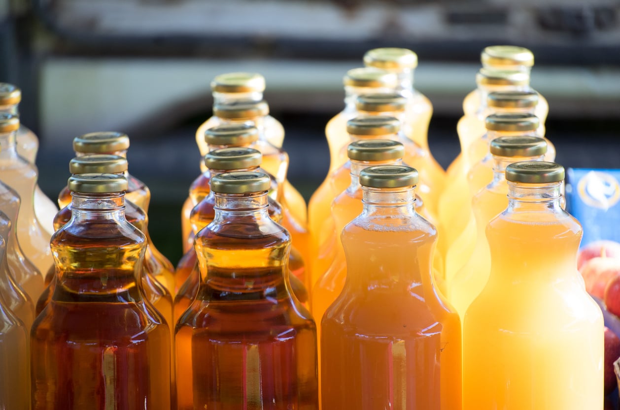 Clear glass bottles filled with various shades of orange and amber liquid, likely juice, are arranged in rows. The bottles have gold caps and sunlight highlights their colors.