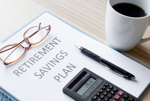 A clipboard with a document titled "Retirement Savings Plan" sits on a desk alongside eyeglasses, a pen, a calculator displaying numbers, and a cup of coffee.