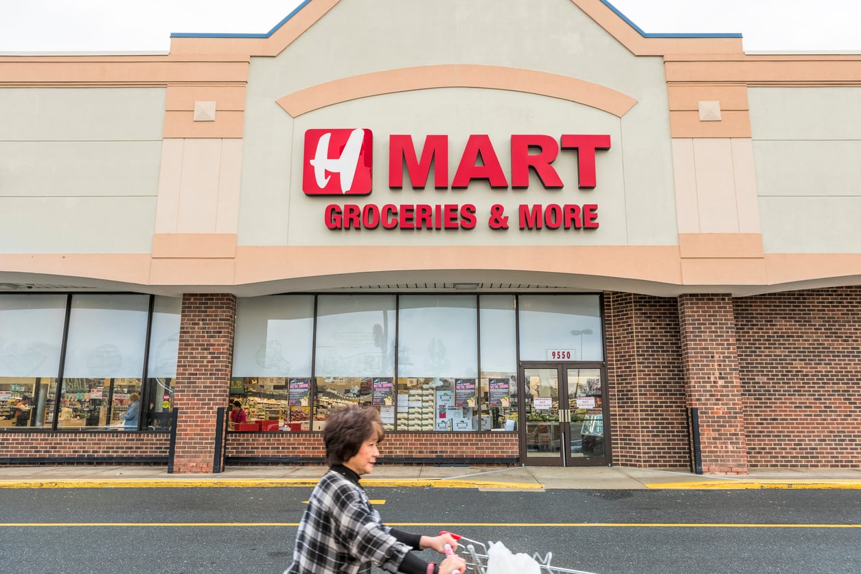 A person pushes a shopping cart in front of an H Mart grocery store with a large red sign that reads "H Mart Groceries & More" on the building's beige and brick exterior.