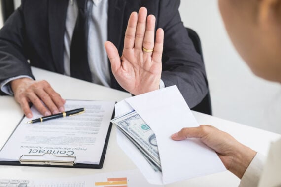 A person in a suit raises a hand to refuse an envelope filled with cash, which is being offered by another person. A contract and pen are visible on the desk, suggesting a business setting.