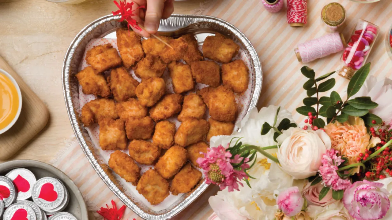 A hand wearing a ring picks up a chicken nugget from a heart-shaped tray filled with nuggets, surrounded by flowers, dipping sauces, and decorative ribbons on a striped tablecloth.