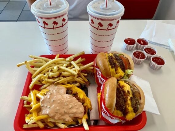 A red tray holds two overrated fast-food burgers, a serving of fries, animal-style fries with melted cheese and sauce, and two drinks in palm tree cups. Four ketchup containers and napkins are beside the tray on a white table.