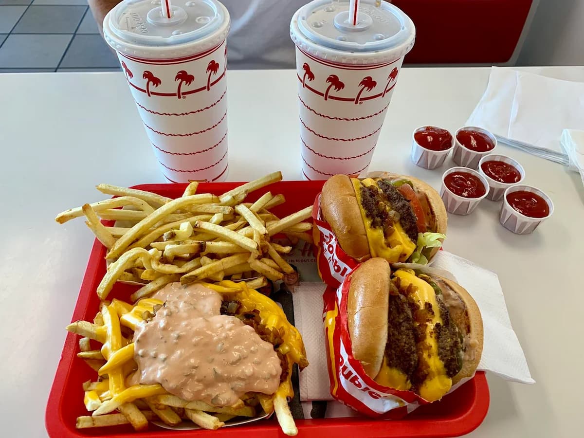 A red tray holds two overrated fast-food burgers, a serving of fries, animal-style fries with melted cheese and sauce, and two drinks in palm tree cups. Four ketchup containers and napkins are beside the tray on a white table.