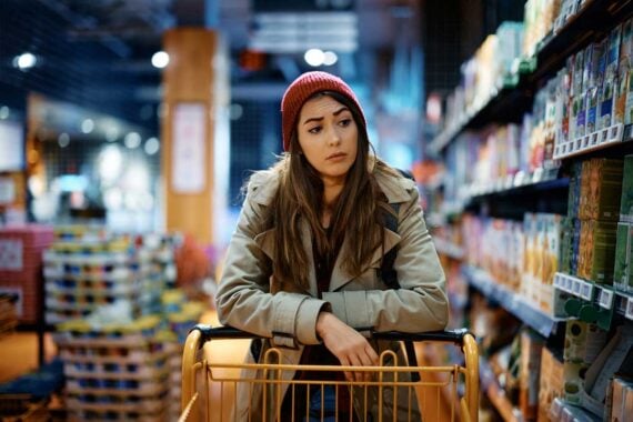 A woman in a red knit hat and beige coat leans on a shopping cart in a grocery store aisle, looking thoughtful or concerned. Shelves of products are visible on both sides of the aisle.