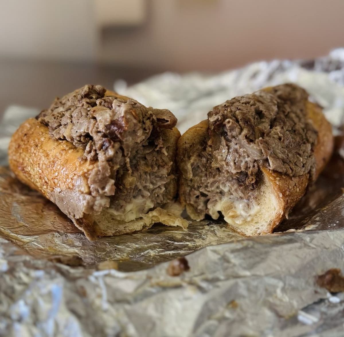 A cheesesteak sandwich cut in half, with chopped beef and melted cheese inside a hoagie roll, sits on crumpled aluminum foil.