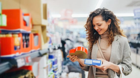 A woman with curly hair smiles while holding and comparing two products in a supermarket aisle, surrounded by brightly colored packaging on the shelves.