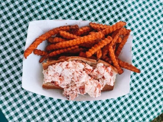 A paper tray with a lobster roll and a serving of sweet potato fries sits on a green and white checkered tablecloth.