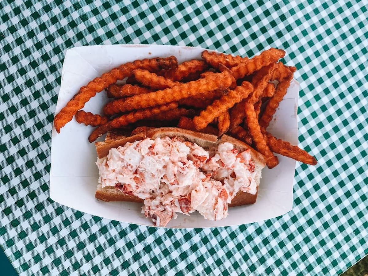 A paper tray with a lobster roll and a serving of sweet potato fries sits on a green and white checkered tablecloth.
