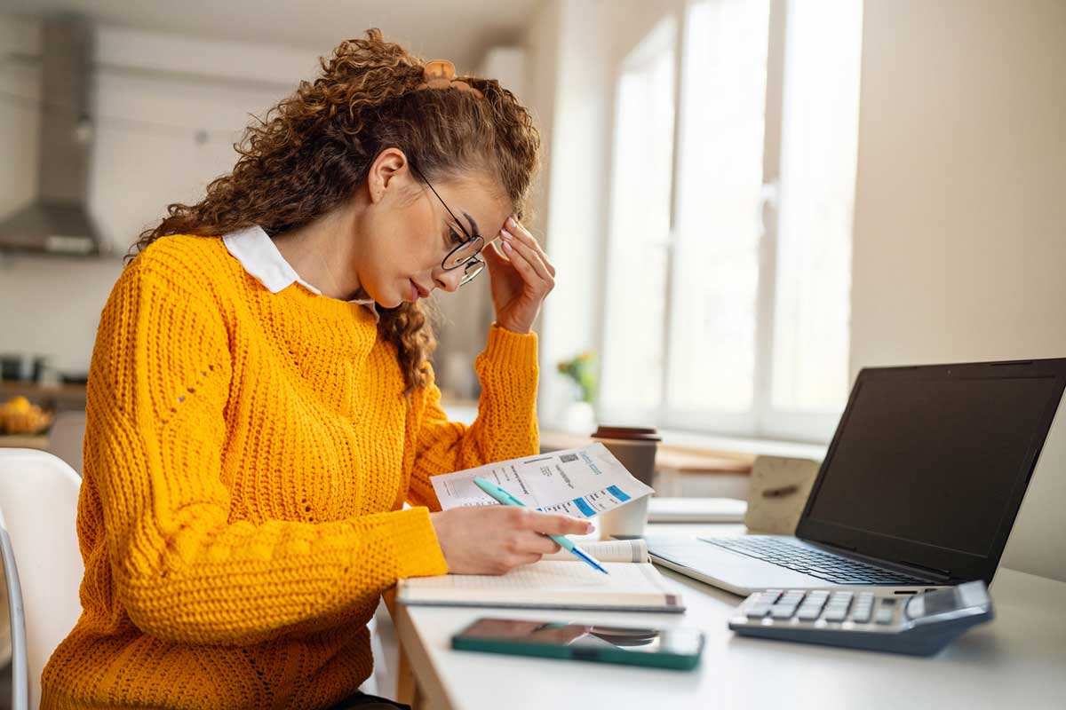 A woman in a yellow sweater sits at a desk, looking stressed while reviewing bills and documents. She has a pen in hand, with a laptop, calculator, and phone nearby in a bright home setting.