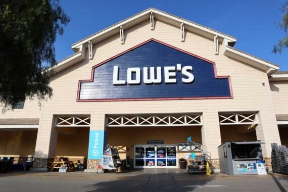 Front view of a Lowe&rsquo;s store entrance with a large blue sign displaying the store&rsquo;s name above the doors. Outdoor merchandise and carts are visible near the entrance amid news of potential 2026 layoffs.