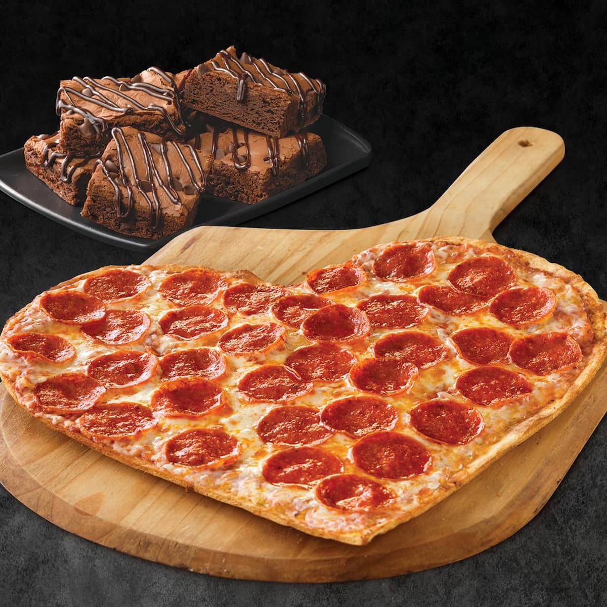 A heart-shaped pepperoni pizza sits on a wooden paddle, with a plate of chocolate-drizzled brownies in the background, all against a dark surface.