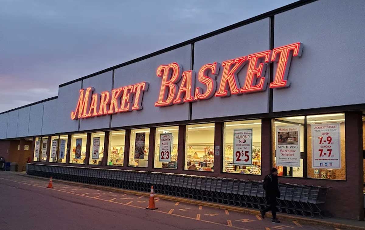 A large Market Basket, one of the top regional grocery chains, stands out at dusk with bright red neon signage and window posters. Shopping carts are lined up outside as a person walks by the entrance.