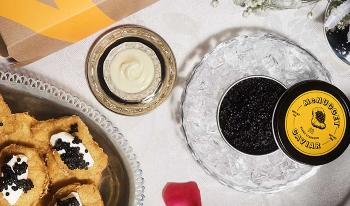 A tin of black caviar on ice, a small dish of creamy sauce, and chicken nuggets topped with sauce and caviar are arranged on a white tablecloth. A takeout box and flower petals are also partially visible.