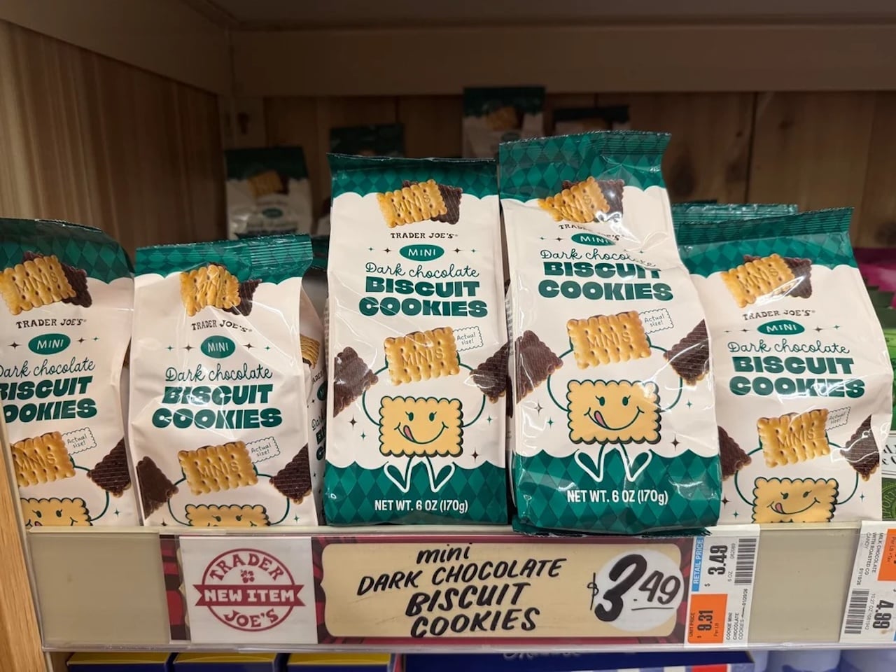 Bags of Trader Joe’s Mini Dark Chocolate Biscuit Cookies are displayed on a store shelf, with a price tag of $3.49 underneath. The packaging is white and green, featuring a smiling cookie illustration.
