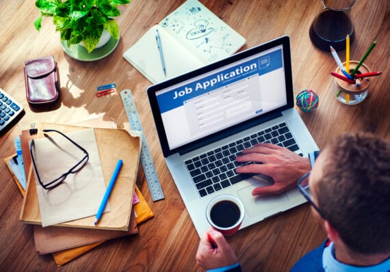 A person holding a cup of coffee sits at a desk cluttered with papers, glasses, and office supplies, while using a laptop displaying an online job application form.