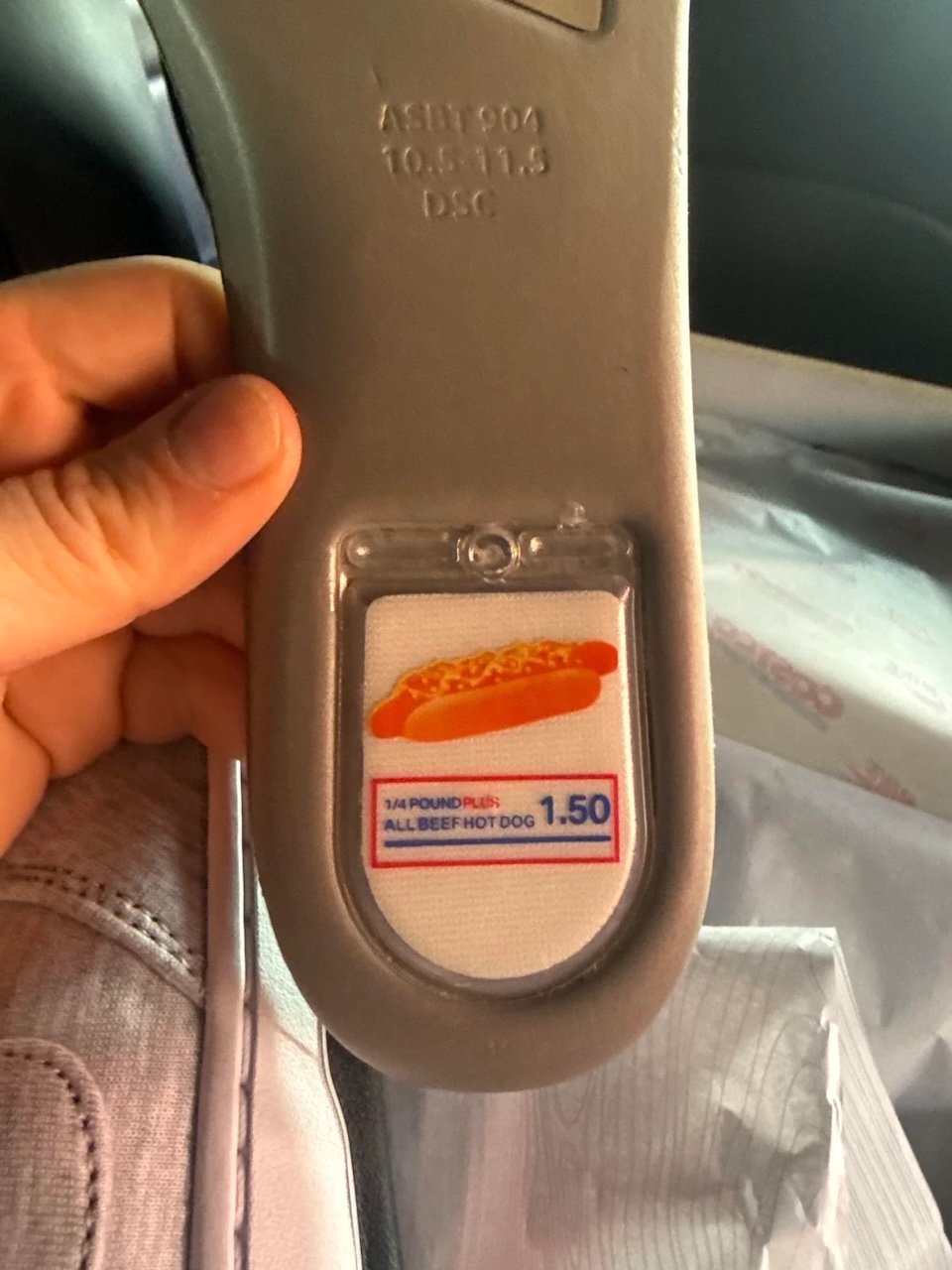 A hand holds a gray plastic handle with a label showing a hot dog and text reading "1/4 POUND PLUS ALL BEEF HOT DOG 1.50". Part of a sneaker and some bags are visible in the background.