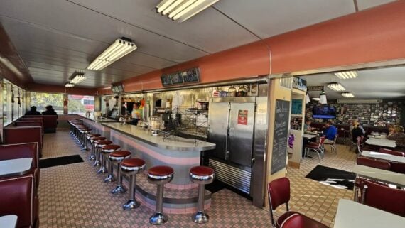 A retro diner with red bar stools lined up at a long counter, checkered tile floor, booth seating, and customers dining in the background. The decor features vintage accents and bright overhead lights.