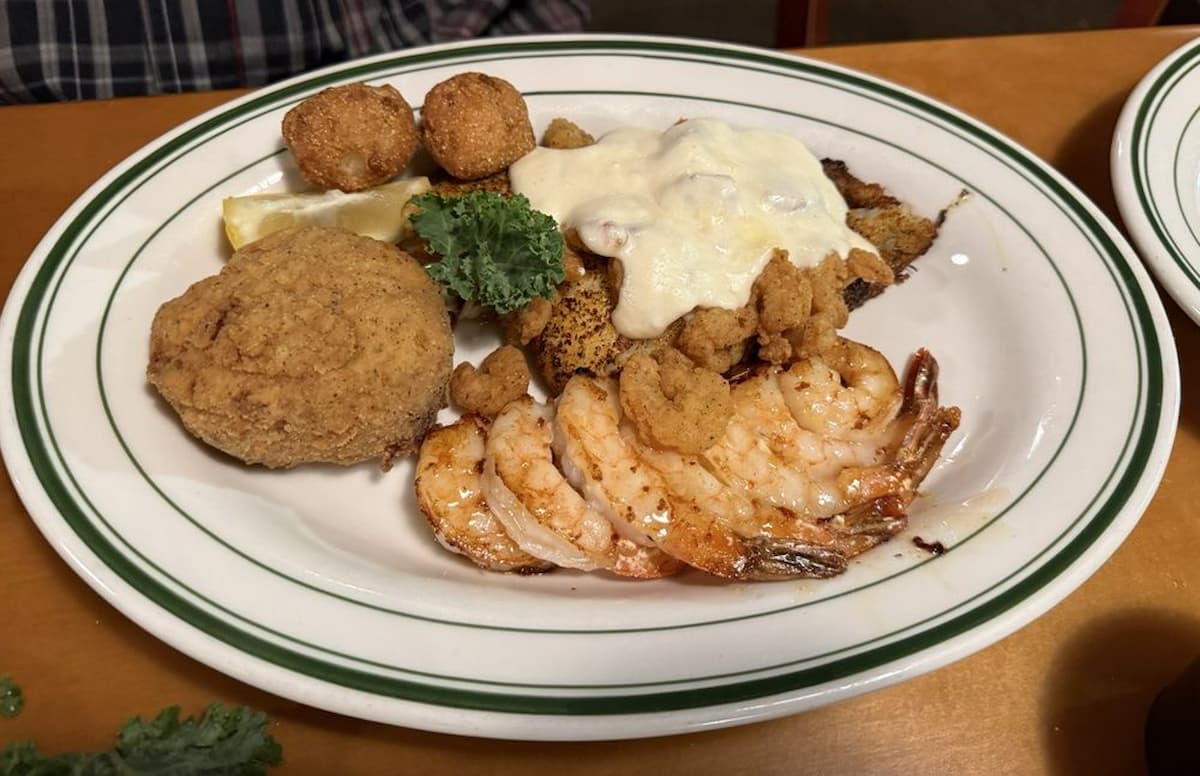 A plate of grilled shrimp, fried hush puppies, a stuffed crab, and a fish fillet topped with creamy sauce, garnished with parsley and a lemon wedge. The food is served on a white plate with green stripes.