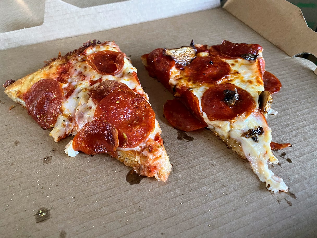 Two slices of pepperoni pizza with melted cheese and a few mushrooms rest in a brown cardboard pizza box. The crust is slightly crispy and the box is mostly empty.