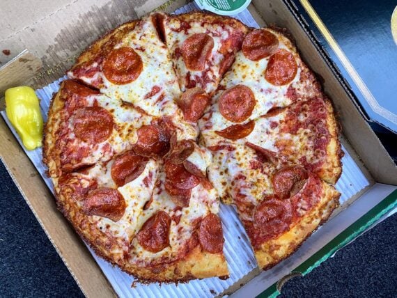 A pepperoni pizza with melted cheese and eight slices sits in an open cardboard box. A yellow pepper and a container of dipping sauce are next to the pizza.
