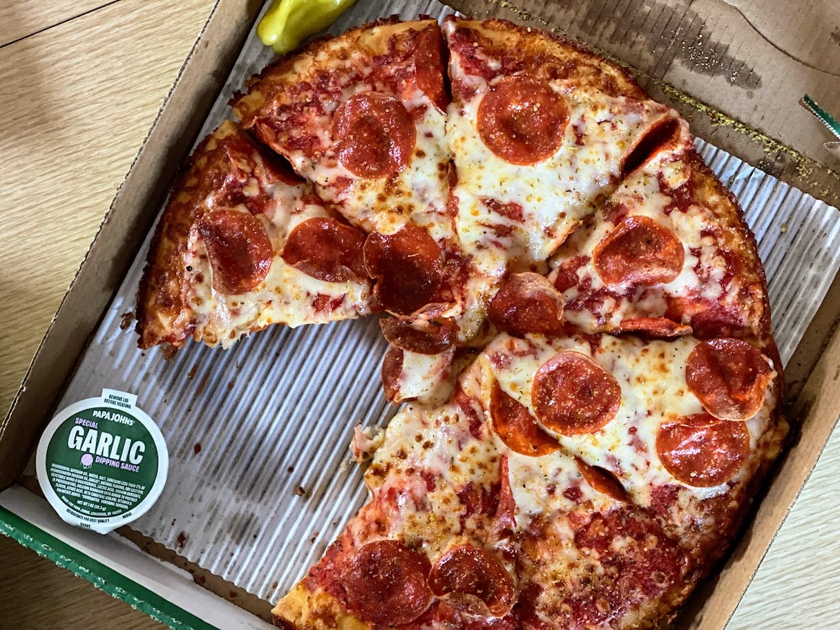 A pizza with pepperoni and melted cheese in an open box, with two slices missing. A container of garlic sauce and a yellow pepper are placed beside the pizza.