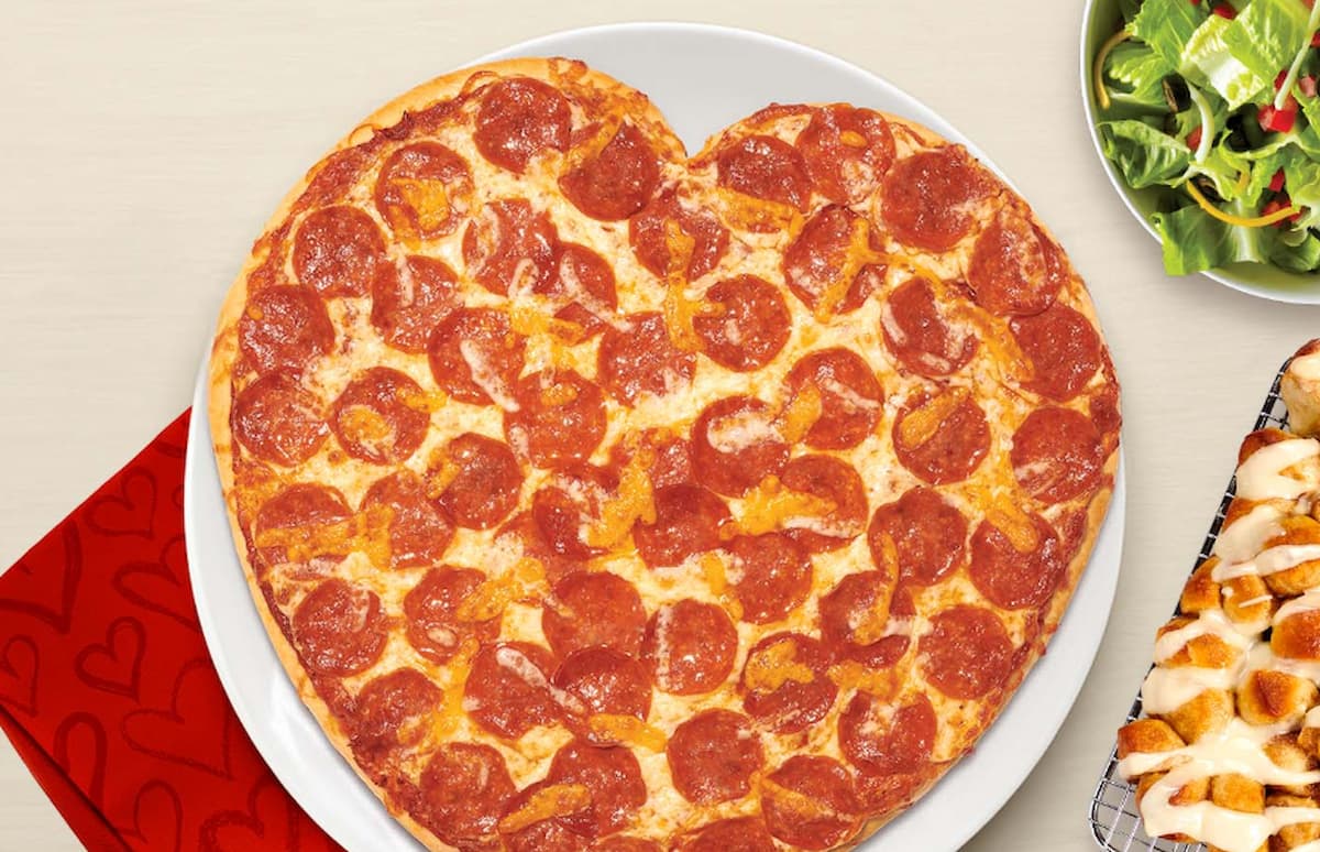 A heart-shaped pepperoni pizza on a white plate, with a red napkin, a bowl of salad, and a tray of dessert rolls nearby on a light-colored table.