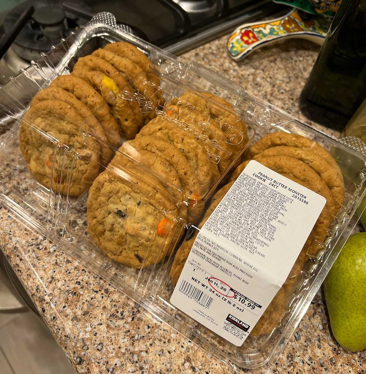 A clear plastic container labeled “Gourmet Butter Monster Cookies with M&M’s,” one of the new Costco items, sits on a speckled kitchen countertop near a green fruit and a gas stove.