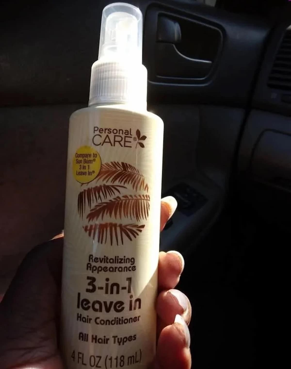 A hand holds a bottle of Personal Care 3-in-1 Leave In Hair Conditioner (4 fl oz, 118 mL) for all hair types inside a car. The bottle has a spray top and a leaf design on the label.
