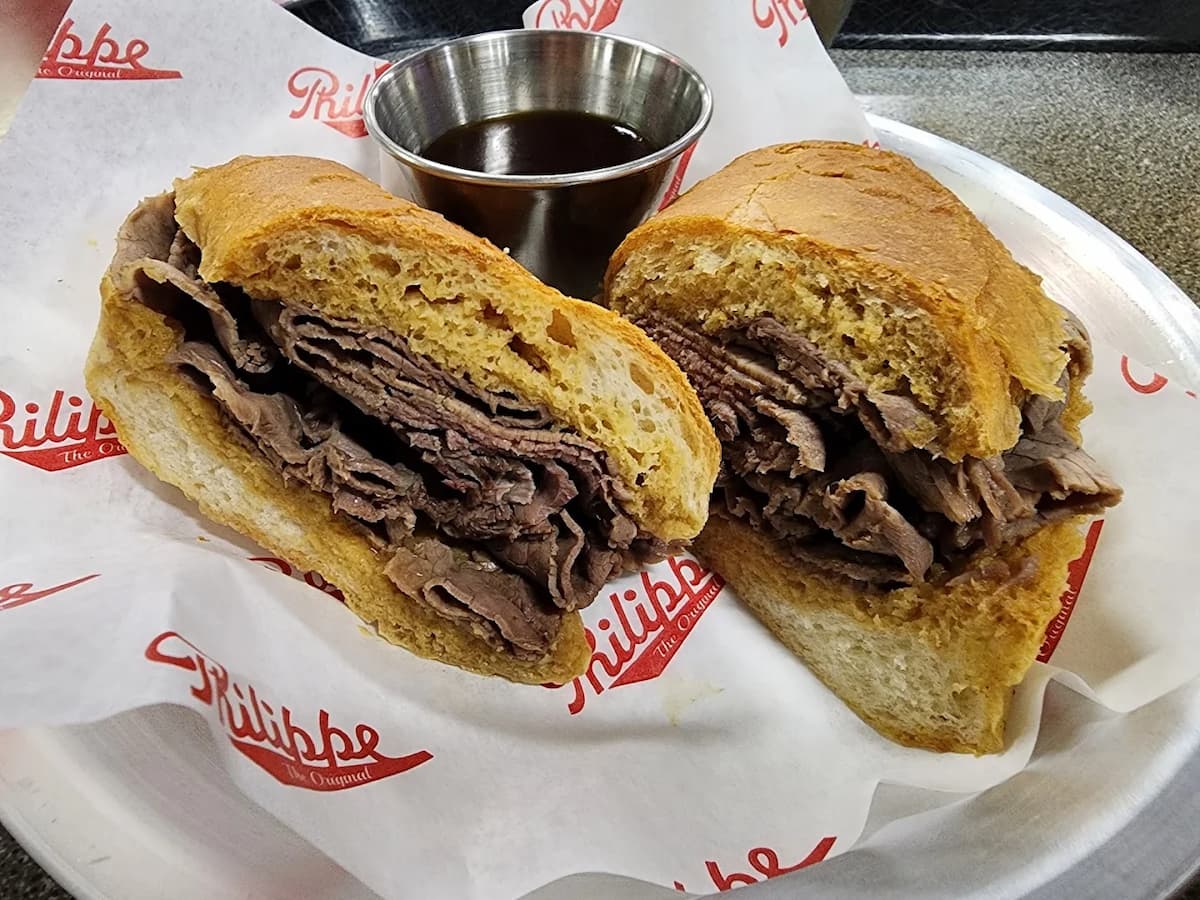 A French dip sandwich cut in half, filled with sliced roast beef on a crusty baguette, served on Philippe-branded paper with a cup of au jus on the side.