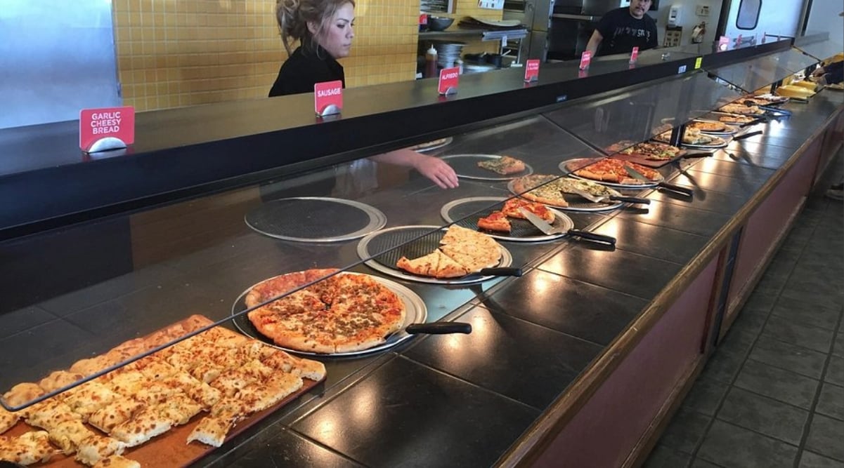 A pizza buffet featuring all-you-can-eat deals under $20 offers various types of pizza and garlic cheesy bread under a glass sneeze guard. A woman serves food behind the counter, while signs label each delicious pizza variety.