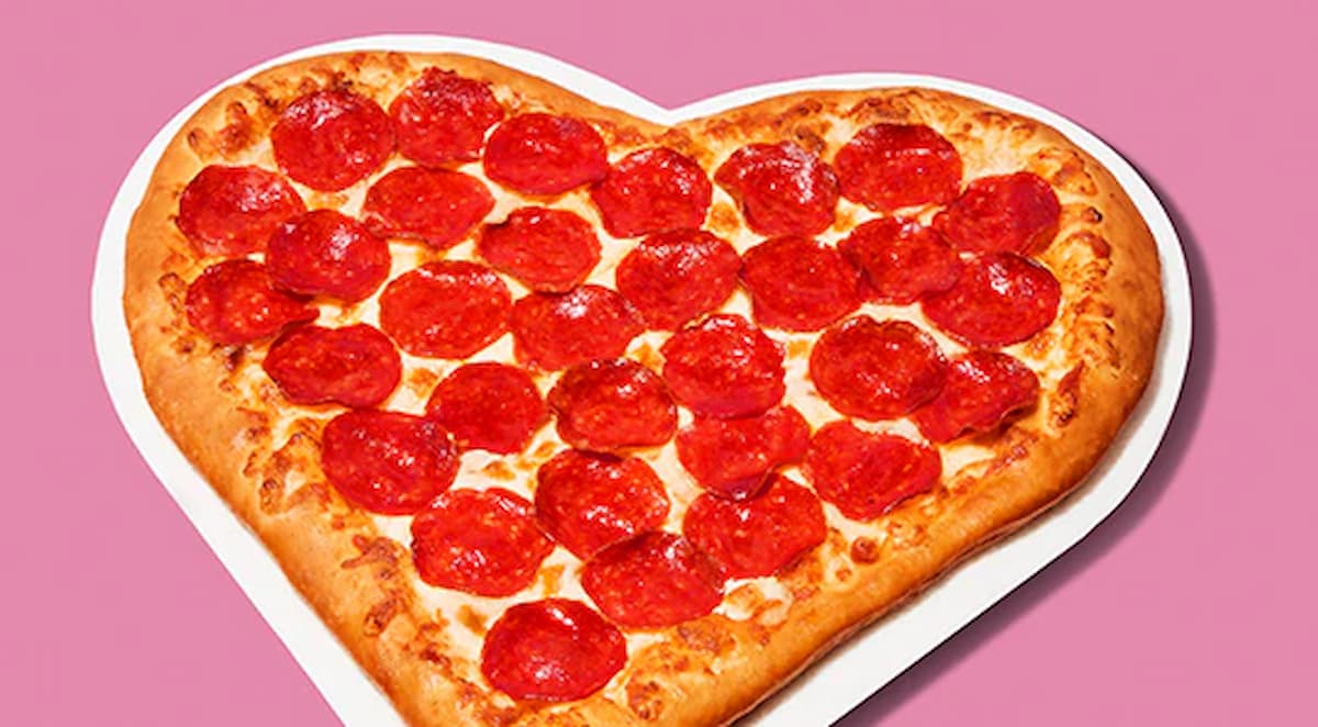 A heart-shaped pepperoni pizza with melted cheese sits on a white plate against a pink background.
