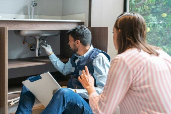 A plumber inspects pipes under a kitchen sink while a woman watches and gestures. The plumber holds a clipboard and wears gloves and a vest. The scene is indoors near a large window with greenery outside.