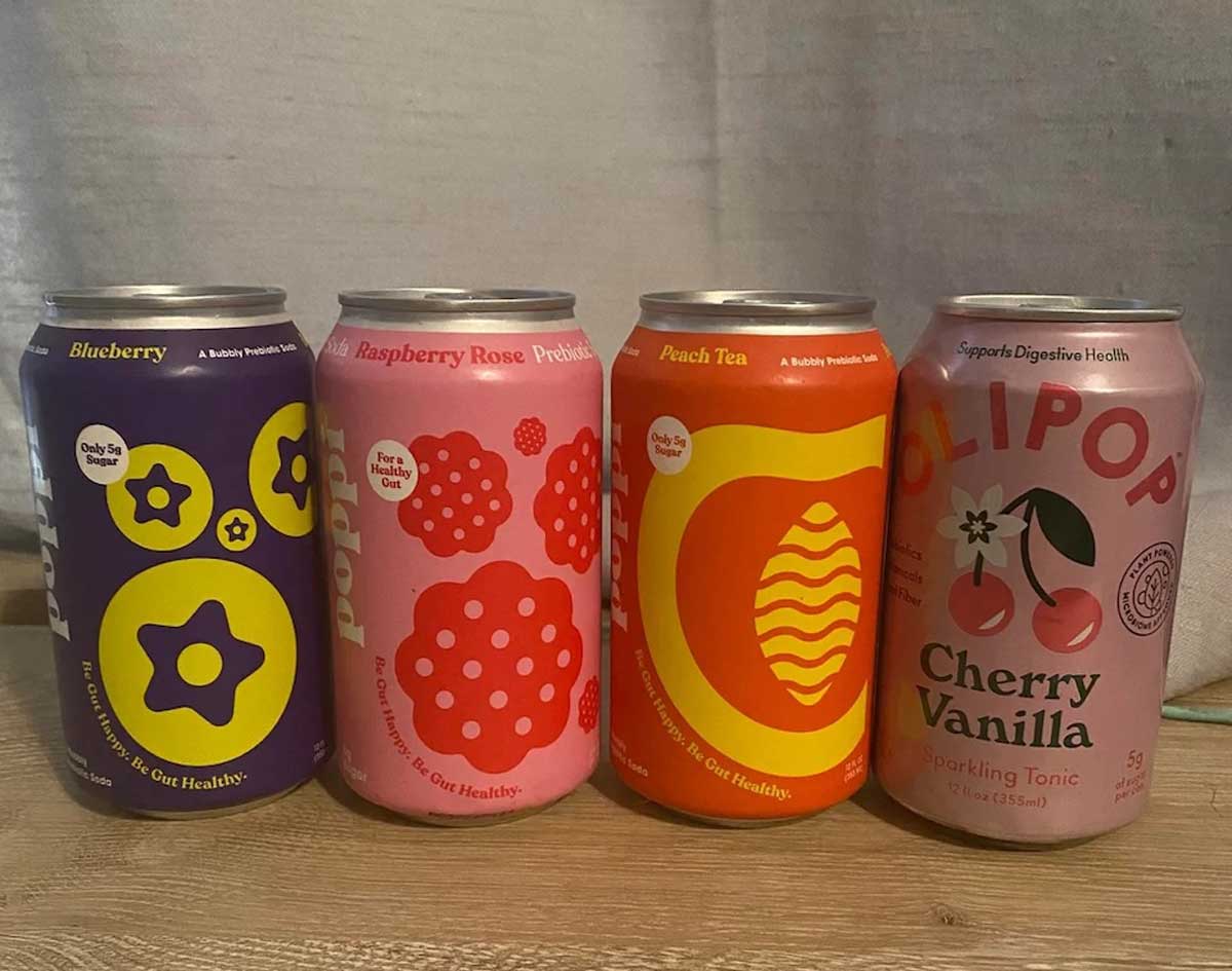 Four colorful cans of OLIPOP sparkling tonic—Blueberry, Raspberry Rose, Peach Tea, and Cherry Vanilla—are displayed side by side on a wooden surface, highlighting them as exciting new Costco items to try.