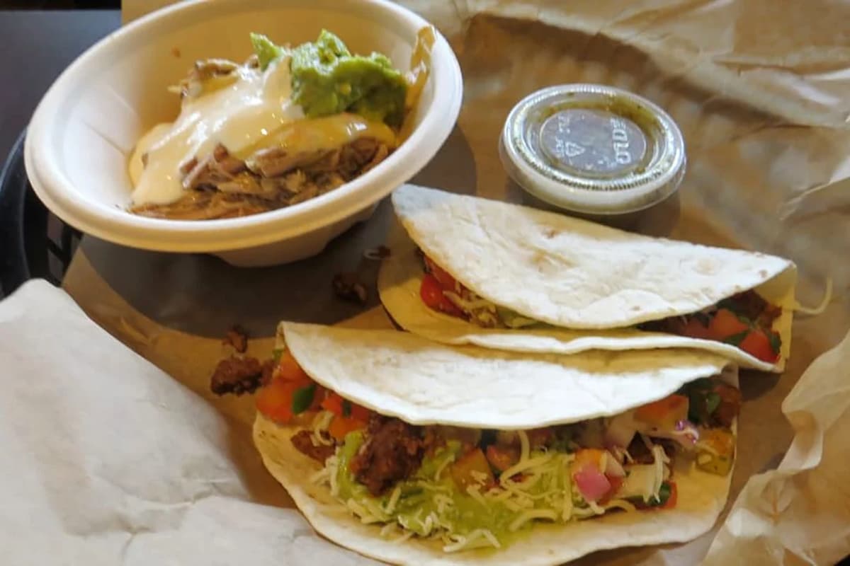 A plate with two soft tacos filled with lettuce, tomato, and meat sits beside a bowl of shredded chicken topped with sauce and guacamole, with a small container of green salsa nearby.