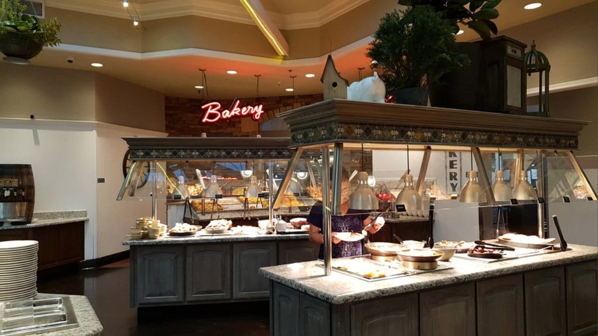 A buffet restaurant interior with all-you-can-eat deals under $20, featuring various food stations, trays under warming lights, stacks of plates, a neon "Bakery" sign in the background, and plants with decorative elements atop the counters.