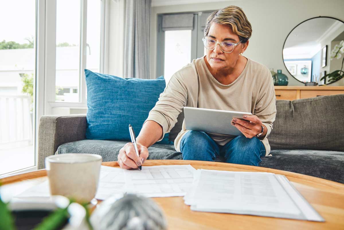 A woman with short hair and glasses sits on a couch, holding a tablet and writing on documents spread out on a coffee table, focused on her work in a bright living room.