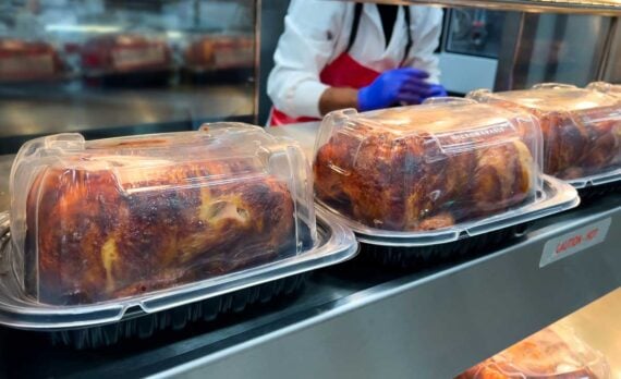 Several rotisserie chickens in clear plastic containers are lined up on a metal counter, with a person wearing a white coat and blue gloves working behind the counter.