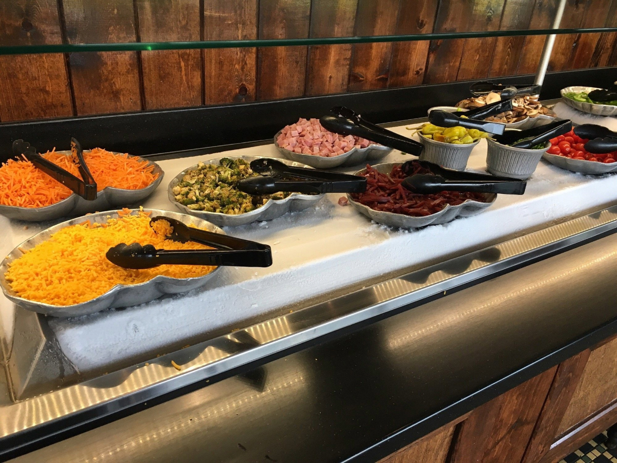 A salad bar with trays of shredded cheese, chopped ham, cherry tomatoes, mushrooms, green peppers, bacon bits, and other fresh toppings—all part of all-you-can-eat deals under $20—each with serving tongs on a bed of ice.