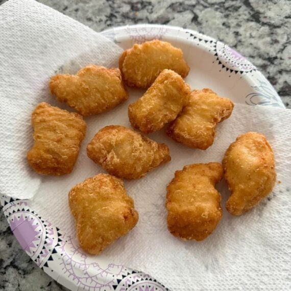 Eight dinosaur-shaped chicken nuggets are arranged on a paper towel atop a decorative paper plate, set on a speckled gray countertop.