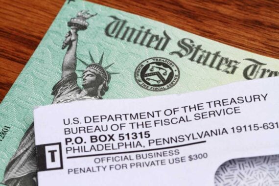 A close-up of a U.S. Treasury check partially covered by an envelope from the U.S. Department of the Treasury, Bureau of the Fiscal Service, on a wooden surface.