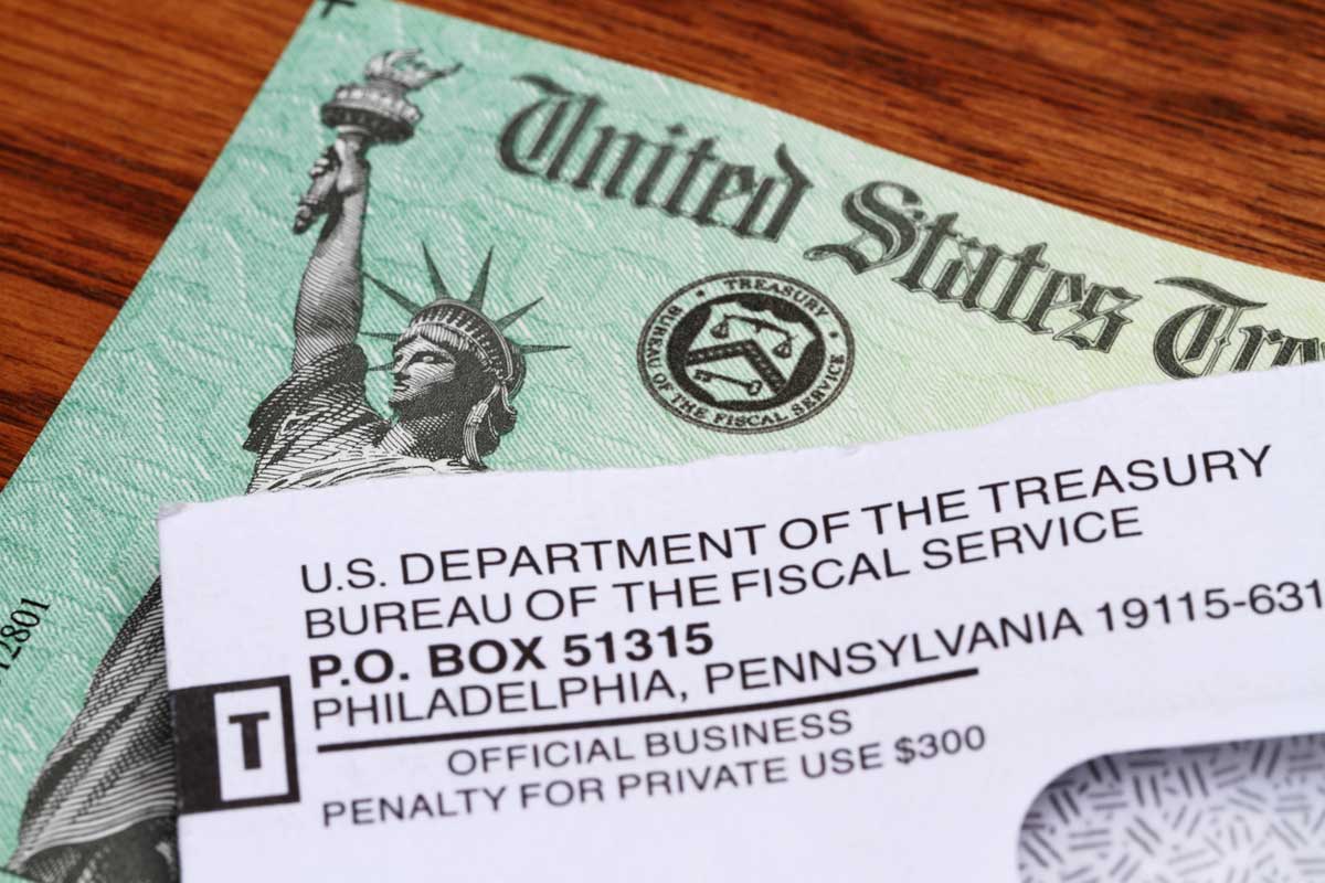 A close-up of a U.S. Treasury check partially covered by an envelope from the U.S. Department of the Treasury, Bureau of the Fiscal Service, on a wooden surface.