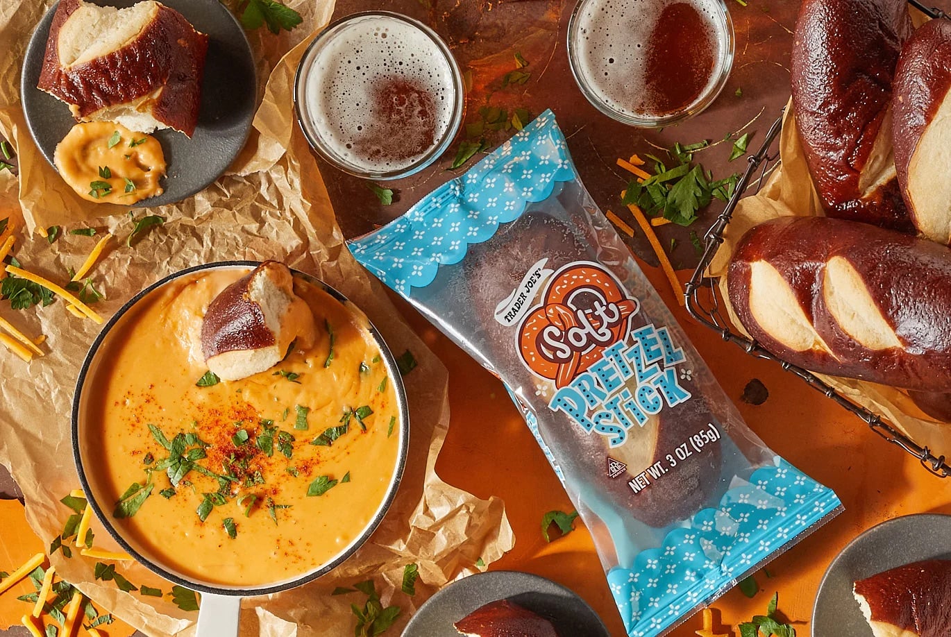 Overhead view of soft pretzel sticks with cheese dip, two glasses of beer, and shredded cheese on a wooden table. A blue bag labeled “Soft Pretzel Stix” is in the center, surrounded by scattered herbs and pretzel pieces.