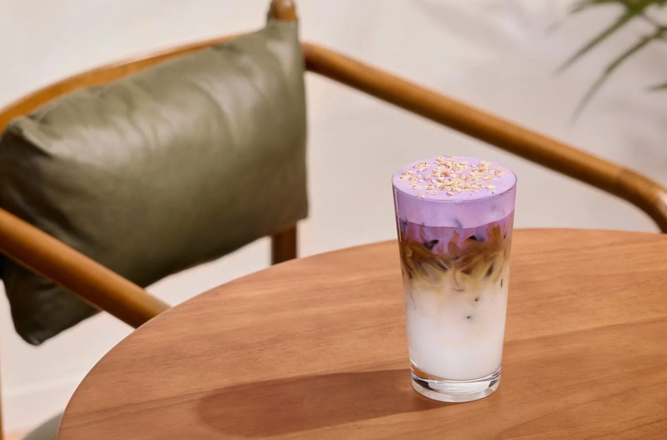 A glass of iced latte with white, brown, and vibrant purple layers topped with flower petals sits on a wooden table next to a chair with a green cushion—an eye-catching treat from the new fast-food menu.
