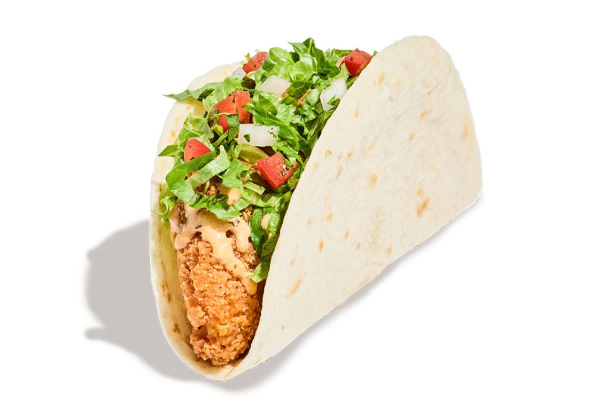 A new fast-food menu item: a soft flour tortilla taco filled with crispy fried chicken, chopped lettuce, diced tomatoes, onions, and creamy sauce, set against a white background.