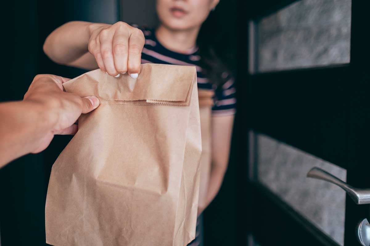 A person hands a brown paper bag to another person standing in a doorway. Only the upper bodies and hands are visible; the recipient wears a striped shirt.