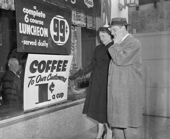 A man and woman in coats smile while looking through a cafe window with signs advertising a 6-course luncheon for 99 cents and coffee for 1 cent a cup. An older man sits inside the cafe, visible through the window.
