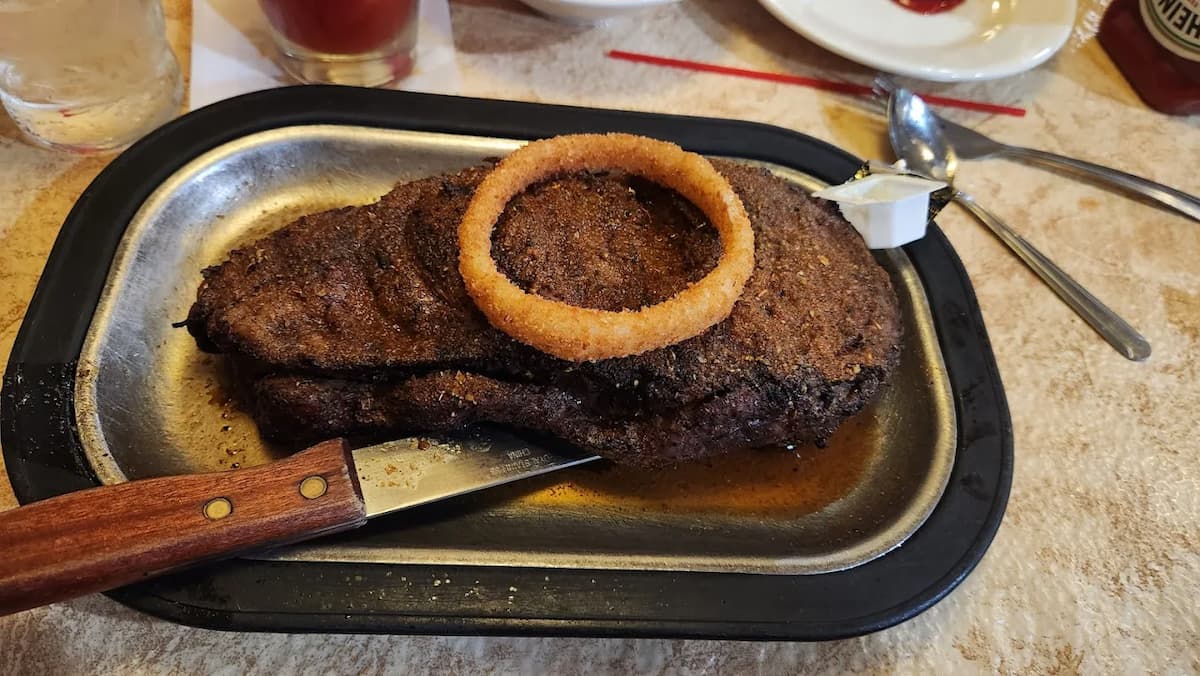 A cooked steak on a metal plate with a wooden-handled knife, topped with a single onion ring and a packet of butter on the side.