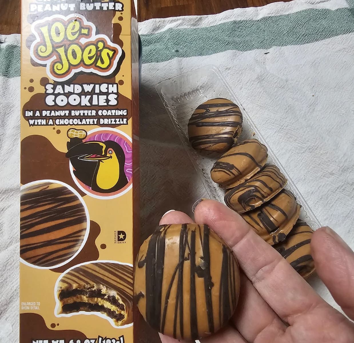 A hand holds a round cookie coated in peanut butter with chocolate drizzle. Next to it is an open package of Trader Joe's Peanut Butter Joe-Joe's sandwich cookies and the product box displaying the cookies and branding.